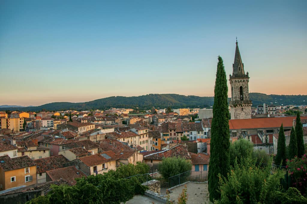 Vue panoramique d'une ville provençale au coucher du soleil, avec toits de tuiles rouges et clocher d'église, dominée par des collines boisées.