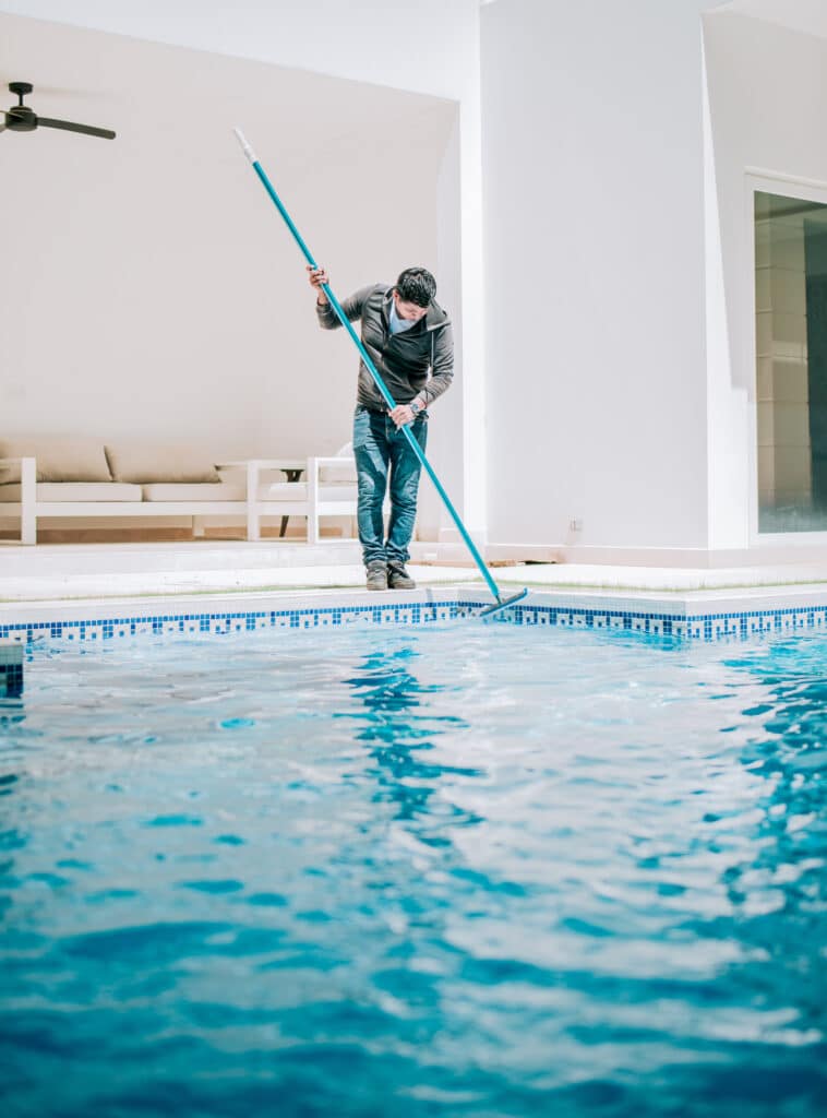 Homme en blouson nettoyant une piscine extérieure avec un balai-brosse, sur fond de murs blancs et de canapé clair.