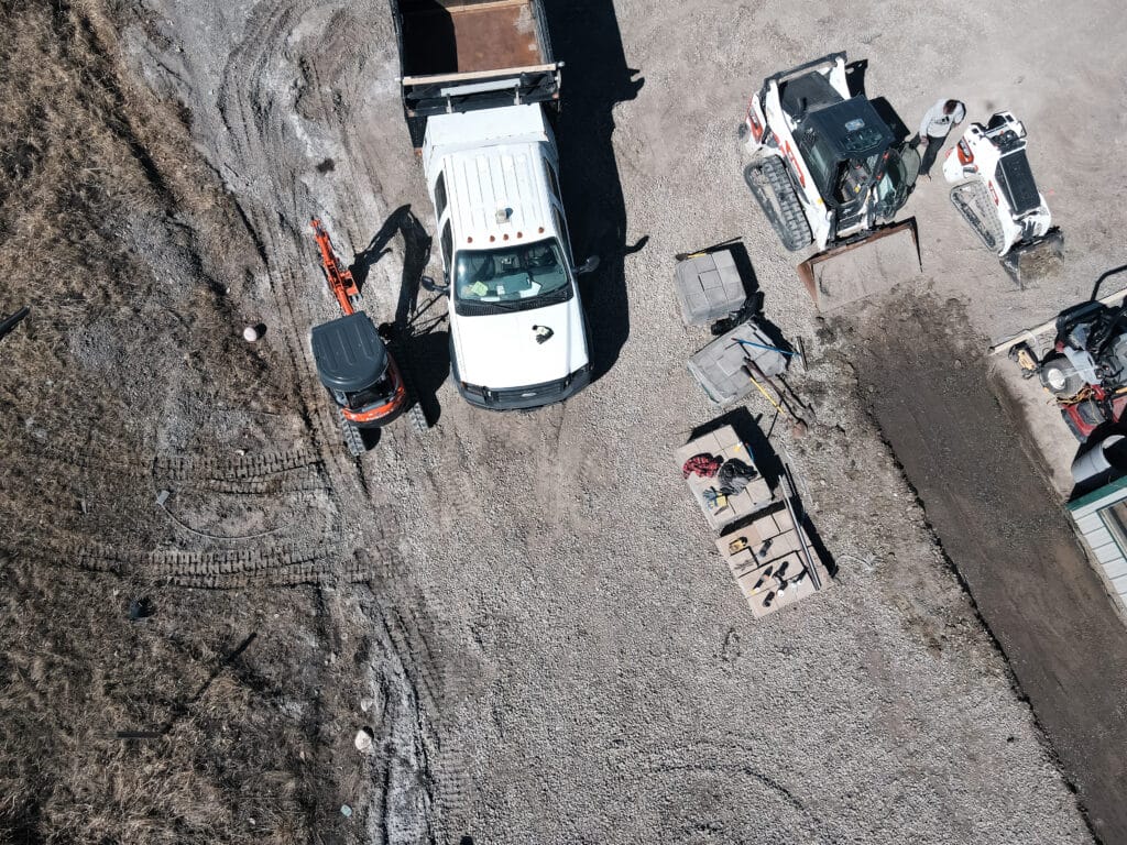 Vue aérienne d'un chantier avec camion blanc, mini-pelle Kubota orange, chargeuses Bobcat, ouvrier et matériaux de pavage.