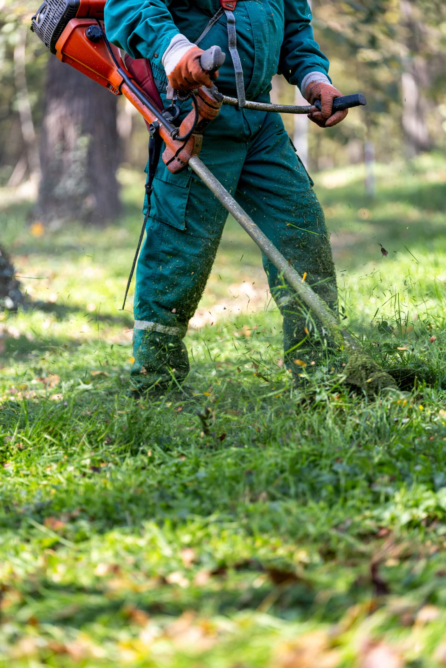 Homme en salopette verte utilisant une débroussailleuse pour couper l'herbe haute, projetant des débris verts.