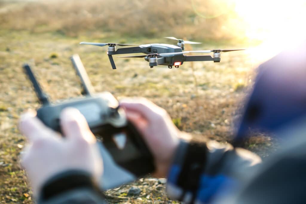 Gros plan sur un pilote manipulant une radiocommande faisant décoller un drone gris à hélices en plein soleil extérieur.