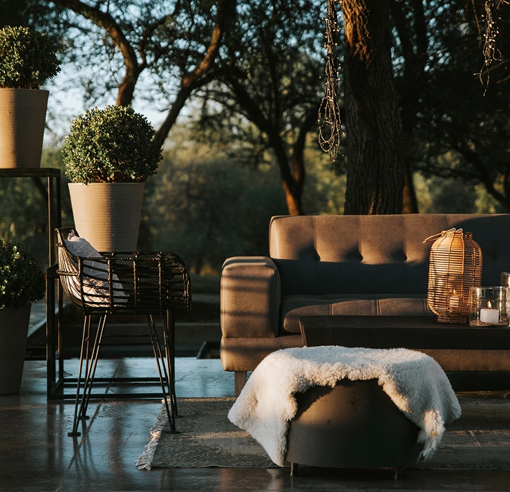 Salon extérieur cosy au crépuscule avec canapé gris, plaid mouton sur pouf, lanterne en osier et plantes en pots.