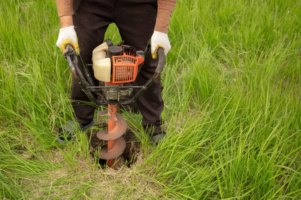 Ouvrier ganté utilisant une tarière thermique pour creuser un trou dans l'herbe verte.