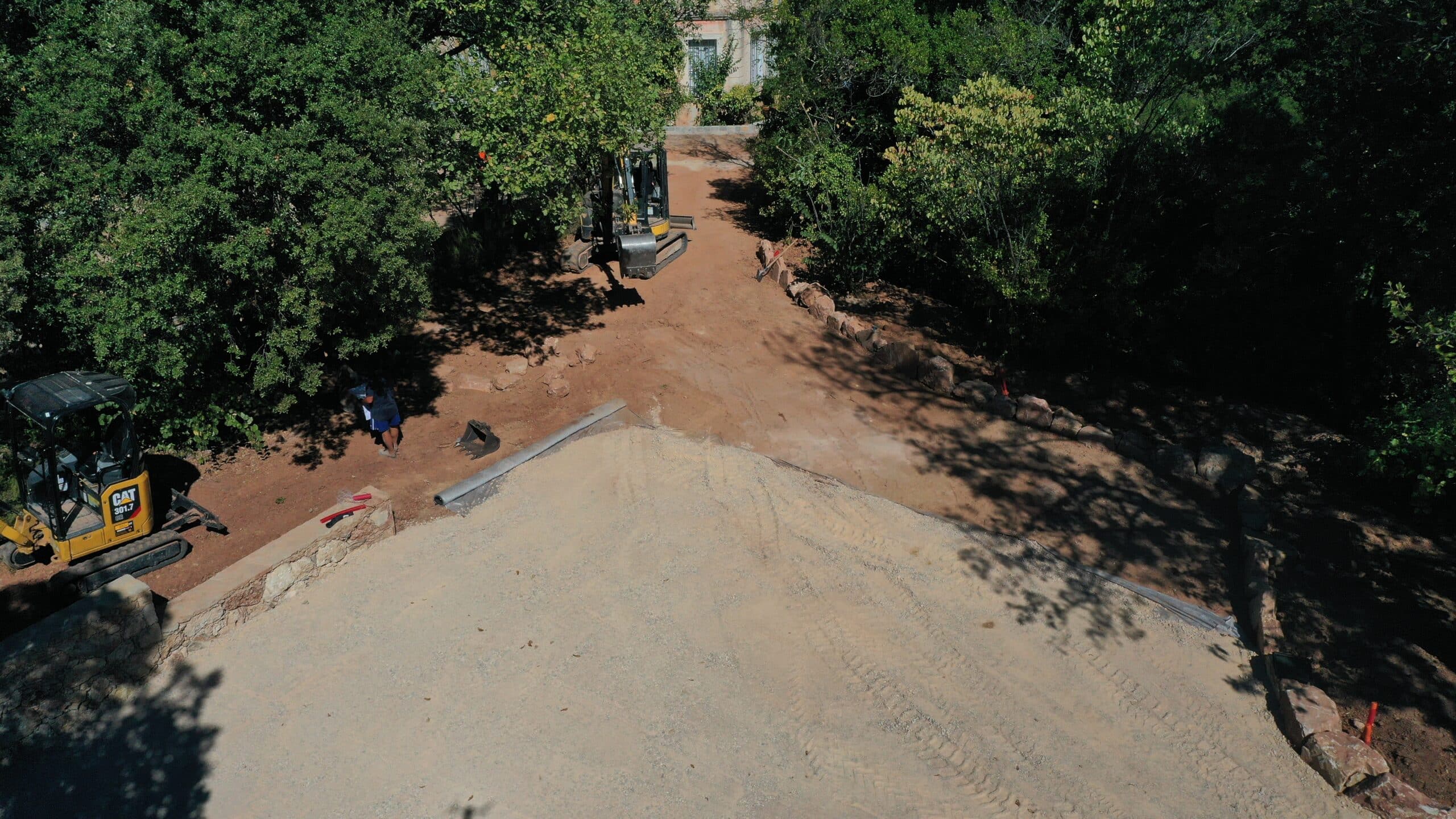 Chantier de terrassement avec mini-pelle CAT 301.7. L'engin nivelle une large allée en gravier, bordée par des roches. Ouvrier au travail.